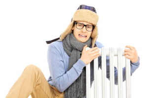 Chilled young man with winter hat sitting next to a radiator isolated on white background